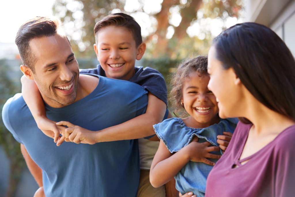 A diverse family of four playfully chats with each other with the son in a piggyback hold from his father and their daughter smiling happily toward her mother. The kids are maybe 8-10 and the parents mid to late 30s.